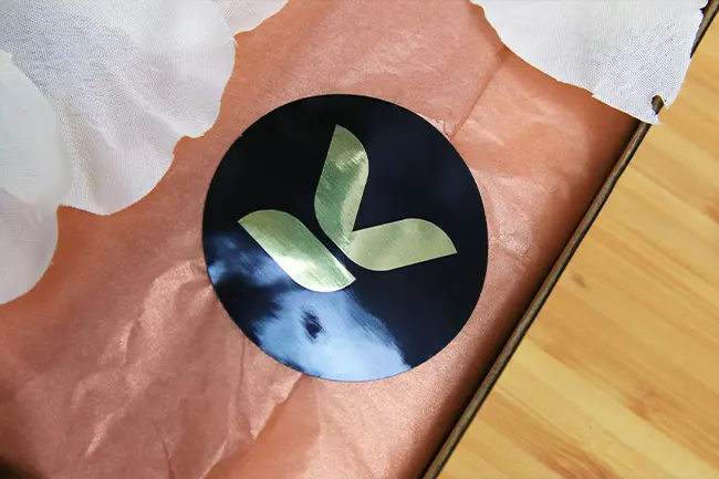 A round black and gold logo sticker sealing peach-colored tissue paper in a box.