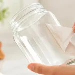 A person struggling to tear off a permanent paper sticker from a clear glass jar, leaving messy paper residue behind.