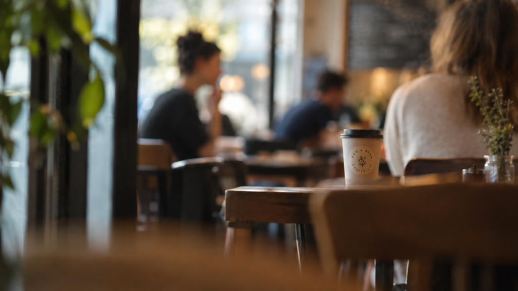 A paper coffee cup with custom logo design sitting on a wooden table inside a cozy cafe.
